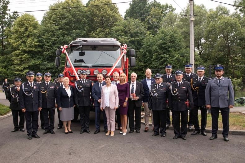 Uczestnicy wydarzenia stoją przed udekorowanym wozem strażackim. W tle widoczna zieleń.