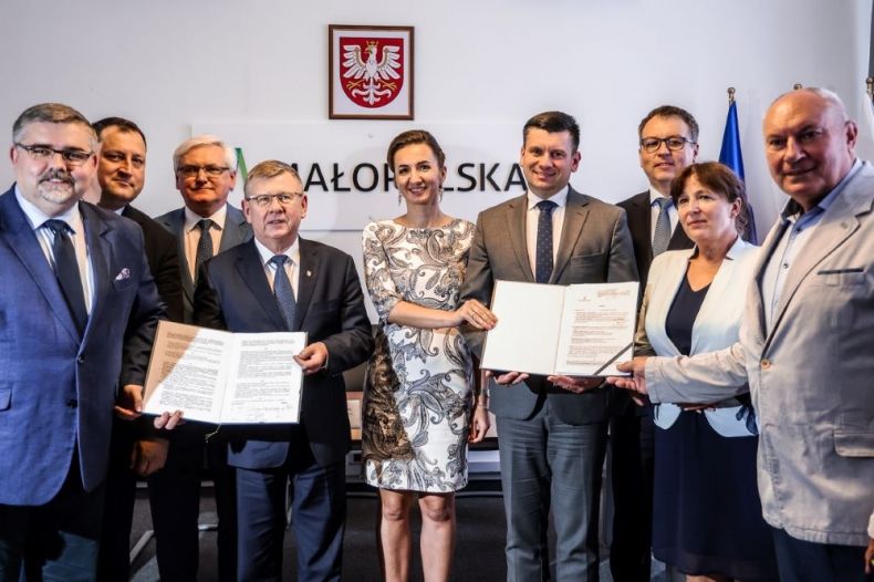 Uczestnicy wydarzenia trzymają podpisane umowy. Z tyłu widoczny herb Małopolski.