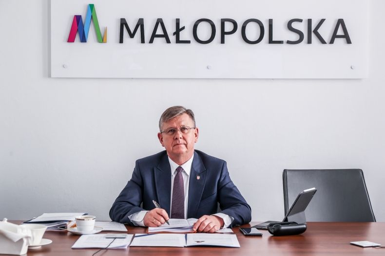 marszałek Witold Kozłowski podczas posiedzenia zarządu, siedzi za biurkiem, na biurku leżą dokumenty i tablet; w tle napis MAŁOPOLSKA