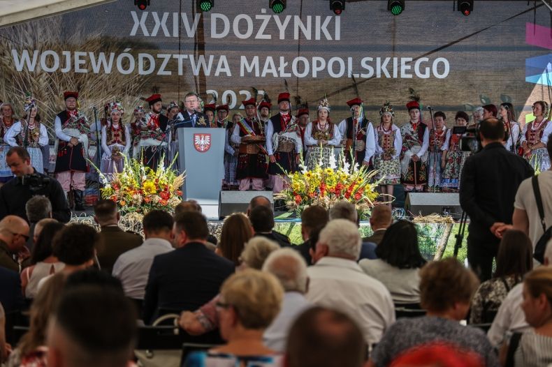 Widok na scenę , na której stoją przemawiający ludzie. Nad sceną widać napis 24 dożynki województwa małopolskiego. W tle widać tłumy ludzi stojących pod sceną. 