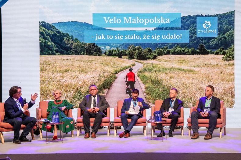 Wszyscy paneliści siedzą na fotelach. Za panelistami widoczny duży slajd z widokiem drogi i pól z napisem Velo małopolska - jak to się stało, że się udało?
