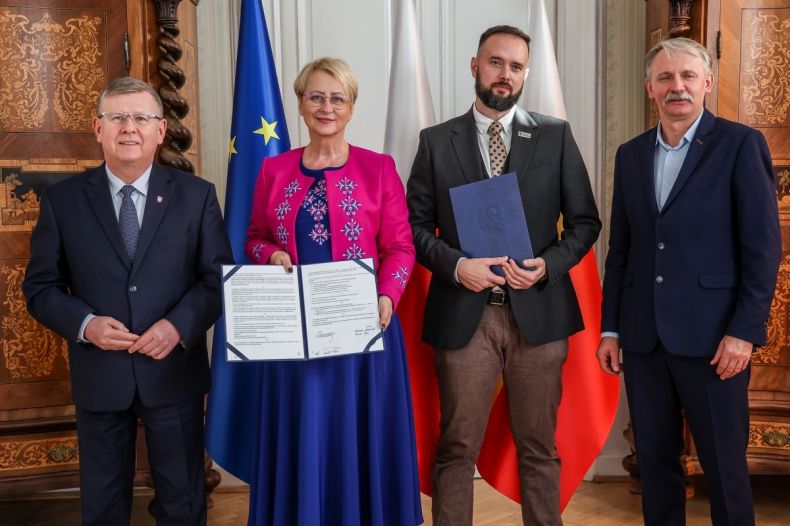 Marszałek Witold Kozłowski, Iwona Gibas z zarządu województwa, Michał Murzyn oraz mężczyzna stoją przy stole i trzymają egzemplarze podpisanych umów. W tle widoczne flagi: UE, Małopolski i Polski.