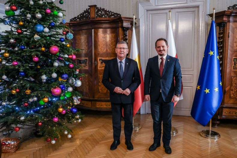 Marszałek Witold Kozłowski stoi wraz z konsulem w gabinecie. W tle widoczna flaga Unii Europejskiej, obok stoi choinka.
