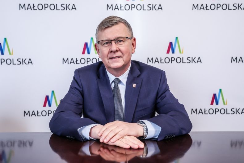 marszałek siedzi za stołem na tle ścianki z logo Małopolski 