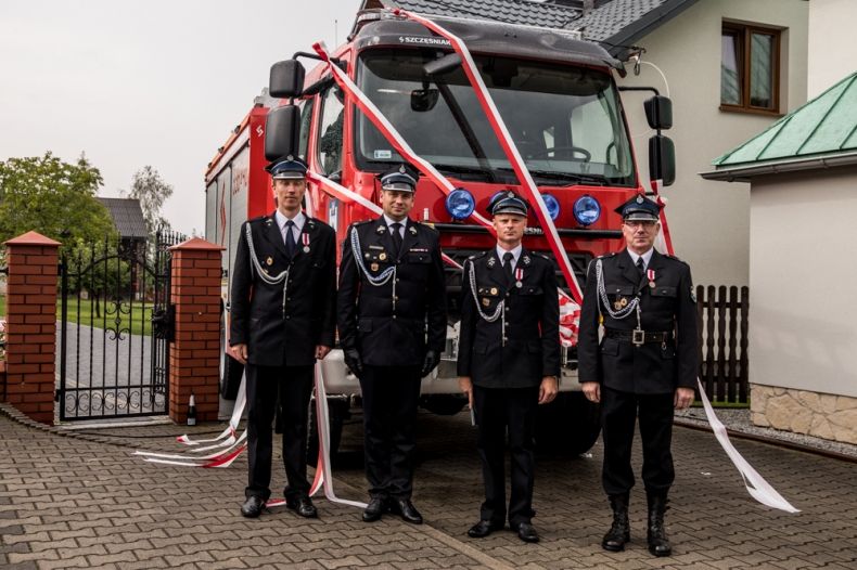 Wicemarszałek Łukasz Smółka i trzech strażaków ochotników w strojach galowych. Mężczyźni stoją w rzędzie na tle czerwonego wozu bojowego. Wóz strażacki ozdobiony jest biało czerwonymi wstęgami. 