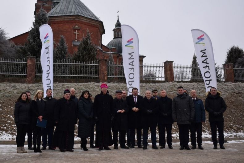 Uczestnicy wydarzenia w Sułoszowej stoją przy drodze. Z tyłu widoczne flagi z napisem Małopolska i zabytkowy kościół.