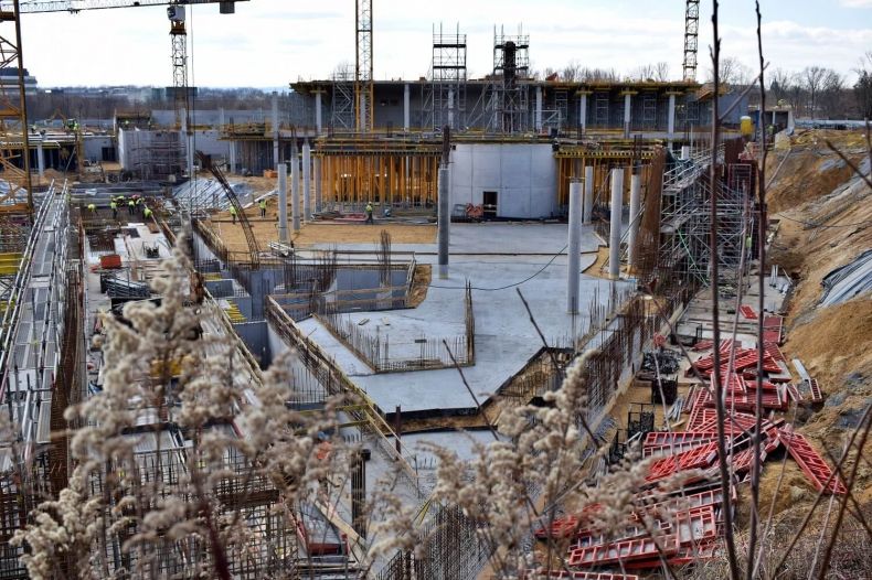 Widok na budowę centrum nauki. Widoczne betonowe wylewki oraz słupy konstrukcyjne oraz trójkątny zarys obiektu.