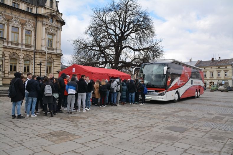 Rynek w Nowym Sączu, kolejka chętnych do oddania krwi ustawiła się do autokaru, w którym oddawana jest krew. Z lewej strony widać część budynku sądeckiego ratusza. Autokar jest w kolorze bieli i czerwieni znajduję się na środku rynku. 