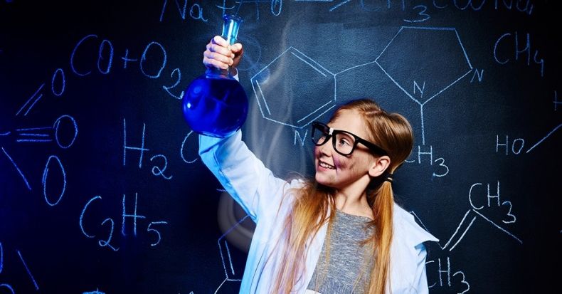 Dziewczynka ubrana w fartuch przygotowuje eksperyment naukowy w butelce, w tle tablica chemiczna