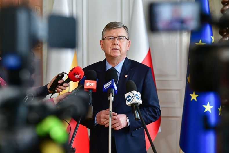 Marszałek Małopolski podczas konferencji prasowej, stoi przed mikrofonami redakcyjnymi, w tle flagi