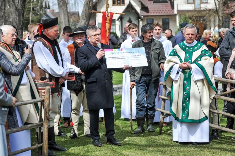 Marszałek Małopolski Witold Kozłowski, stoi na placu przy kościele. Wokół niego widać Baców ubranych w tradycyjne stroje ludowe. Z lewej strony widać księdza proboszcza. Marszałek trzyma wręku symboliczny czek. 