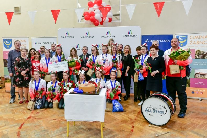 Wnętrze pomieszczenia, cheerleadereki ustawiły się w dwóch rzędach na tle białej ścianki z logami w kształcie litery M i napisem Małopolska. Przednimi ustawiono biały stolik z tortem. Z lewej strony widać grupę ludzi trzymających między innymi kwiaty.