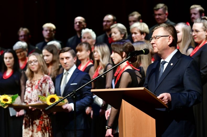Marszałek Małopolski Witold Kozłowski, stoi na tle chóru. Marszałek Stoi perzy brązowym pulpicie - jest w trakcie przemowy i składania gratulacji. 