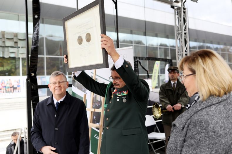 Mężczyzna unosi w górę w geście triumfu, Medal Polonia Minor. Po jego prawej stronie stoi Marszałek Witold Kozłowski po lewej radna Marta Mordarska. 