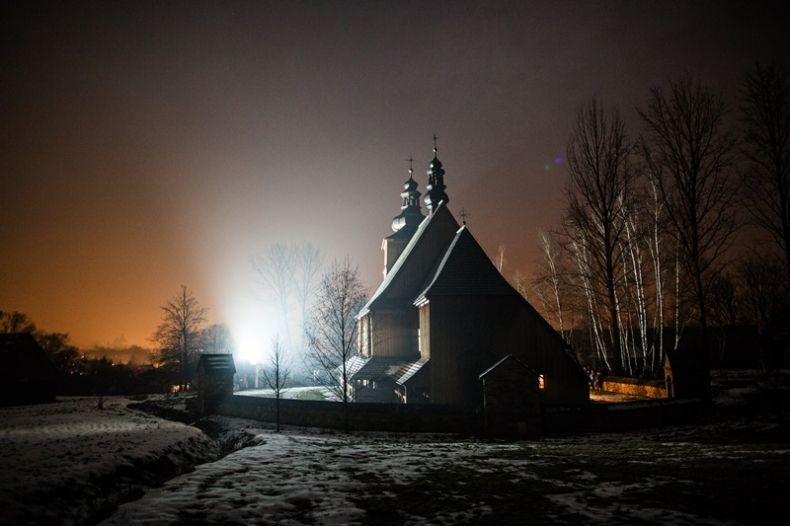 Zdjęcie przedstawiające drewniany kościół w skansenie, nocą w śnieżnej aurze. 