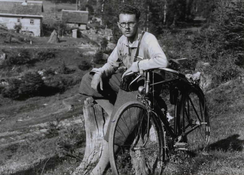 Fotografia archiwalna, czarno - biała a na niej młody Roman Reinfuss przy swoim rowerze, w tle wioska