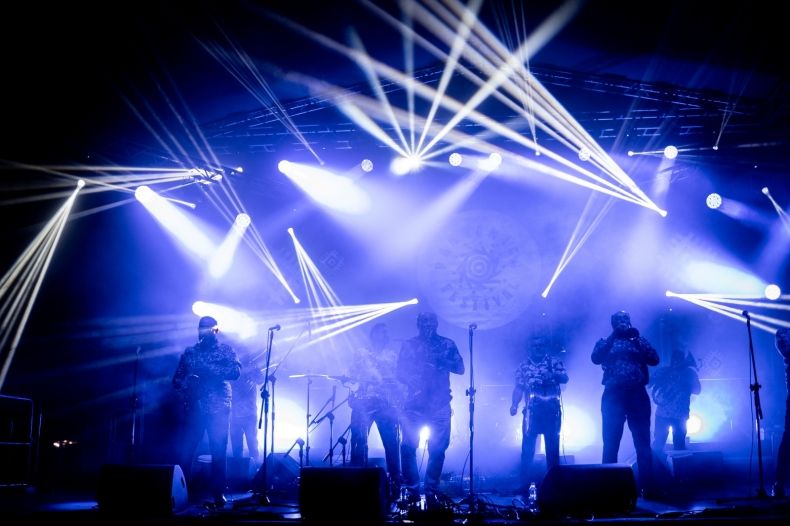 Widok na scenę oświetlona kilkunastoma jasnoniebieskimi snopami światła. NA scenie widać jedynie ciemne sylwetki stojących na niej muzyków. 