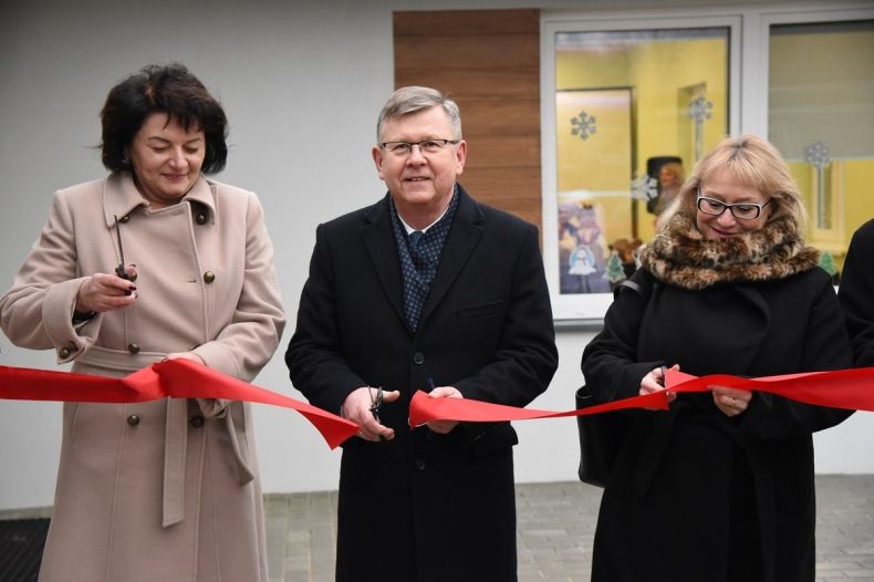 Radna wojewódzka Jadwiga Wójtowicz, marszałek małopolski Witold Kozłowski, wójt gminy Sękowa Małgorzat Małuch, przecinają czerwoną wstęgę przy wejściu do nowego przedszkola w Sękowej. W tyle widać elewację budynku i okna przedszkola.