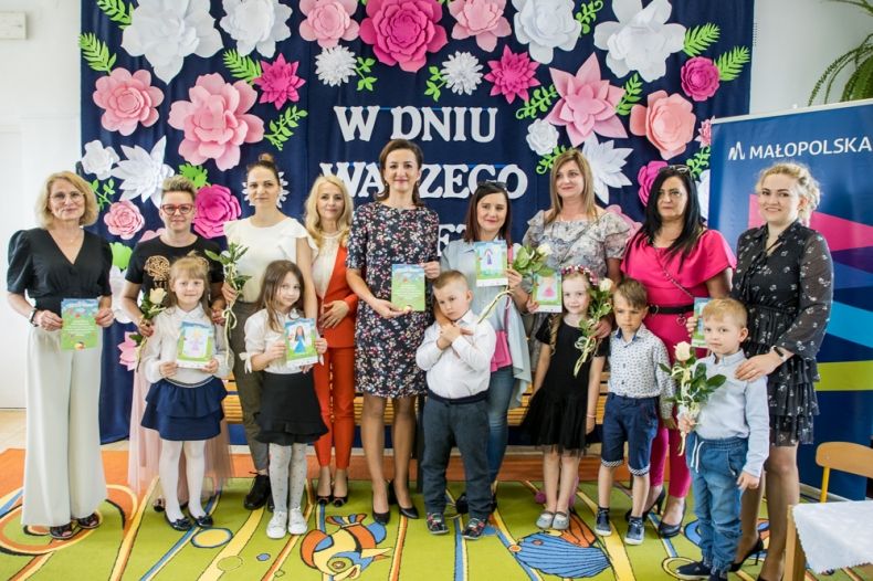 Marta Malec-Lech z zarządu województwa, Beata Kania dyrektor biblioteki, Wioletta Dyngosz-Sapeta dyrektor przedszkola, wszystkie mamy oraz dzieci stoją w przedszkolnej sali. W tle widoczne kwiaty.