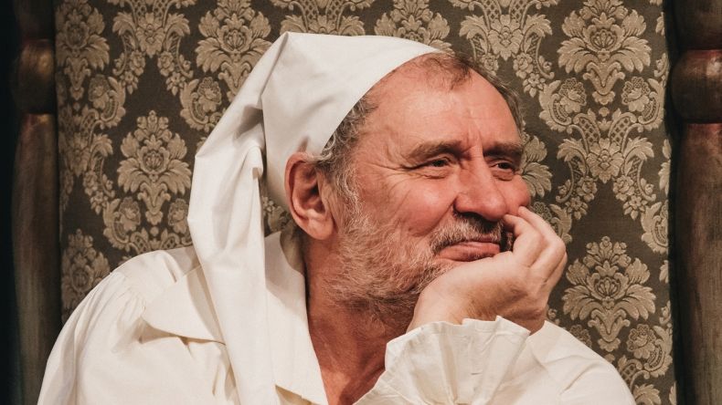 na zdjęciu aktor w białej, spiczastej czapce. Ma na sobie białą koszulę. Podpiera głowę dłonią. Rozmarzony patrzy przed siebie.