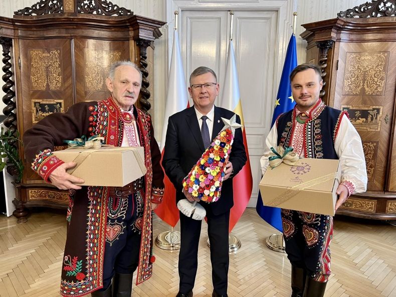 marszałek Witold Kozłowski w towarzystwie dwóch mężczyzn w strojach regionalnych, stoją na tle flag trzymając w ręce prezenty.
