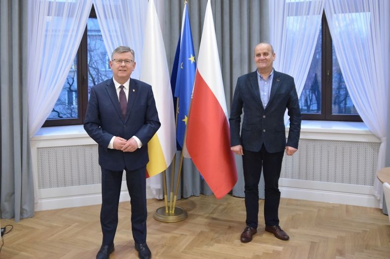 Marszałek Małopolski Witold Kozłowski w agendzie zamiejscowej UMWM w Nowym Sączu i Stanisław Banach Wójt gminy Podegrodzie. Mężczyźni stoją na tle okien i flag Polski, Unii Europejskiej i Małopolski.