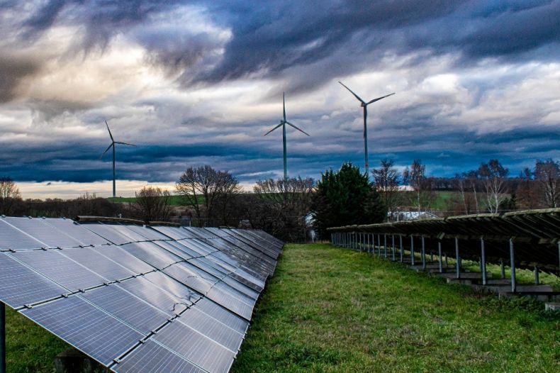 Panele fotowoltaiczne stoją na polu porośniętym trawą, w oddali widoczna instalacja elektrowni wiatrowej