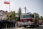 Wóz strażacki jedzie obok strażaków i flagi Polski na maszcie.