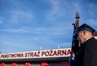 Strażak stoi ze sztandarem przed budynkiem z napisem Państwowa Straż Pożarna.