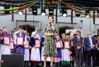 Marta Malec-Lech mówi do mikrofonu, stojąc na scenie, w tle grupa osób w strojach regionalnych