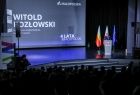 Marszałek Małopolski stoi na scenie przy szarej mównicy. W tle za przemawiającym marszałkiem widać trzy flagi