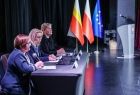 Widok od prawego boku na trzy specjalistki do spraw środków unijnych. Kobiety siedzą przy stole ustawionym na scenie. W tle widać mównice oraz trzy flagi. 