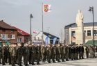 Grupa żołnierzy stoi na płycie rynku, w tle zabudowania miejskie