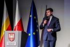 Wicemarszałek Łukasz Smółka w trakcie wystąpienia. Wicemarszałek stoi na scenie w tle widać trzy flagi, polską małopolską i unijną. 