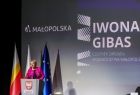 Wicemarszałek Iwona Gibas stoi przy szarej mównicy w tle widać trzy flagi polski unii i małopolski. Po prawej stronie wyświetlono duży napis Iwona Gibas i logo małopolski. 