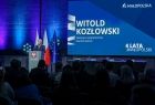 Widok z oddali na scenę , na której ustawioną szara mównice, w tle widać wyświetlony napis. 