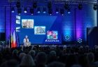 Wicemarszałek Iwona Gibas stoi na scenie przy mównicy, za wicemarszałek widać niebieski ekran multimedialny na którym wyświetlone są zdjęcia.