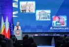 Wicemarszałek Iwona Gibas stoi na scenie przy mównicy, za wicemarszałek widać niebieski ekran multimedialny na którym wyświetlone są zdjęcia.