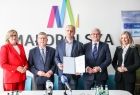 Mężczyzna w kraciastym garniturze prezentuje umowę. Obok marszałkowie i panie poseł.