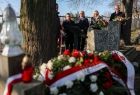 Widok na wieniec złożony a grobie poległych żołnierzy. Kwiaty oraz szarfa wieńca są w kolorach narodowych. W tle widać uczestników mszy żałobnej. 