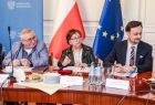 Dwaj mężczyźni i kobieta siedzą. W tle widoczne flagi Polski i UE.