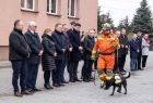 Wicemarszałek Łukasz Smółka stoi z innymi uczestnikami wydarzenia na placu przed remizą. Obok przechodzi ratownik z psem.