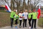 Seniorzy w strojach sportowych stoją na tle krzaków trzymając w rękach flagi biało-czerwone.