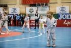 Dziewczynka w stroju karate stoi na hali sportowej. W tle inne dzieci.