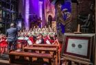 Na pierwszym planie stoi w kościele medal Polonia Minor, z tyłu widoczny dyrygent i zespół "Mazowsze" w strojach ludowych.