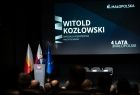 Widok z publiczność na scenę na której stoi Marszałek Witold Kozłowski. W tle za nim wyświetlony jest slajd z jego imieniem i nazwiskiem,. 
