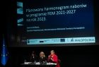 Widok na stół przy który siedzą trzy kobiety. Stół ustawiony jest na scenie, za nim wyświetlają się slajdy z informacjami dotyczącymi Funduszy UE. 