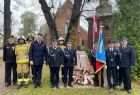 Wicemarszałek Łukasz Smółka stoi ze strażakami przed drewnianym kościołem.