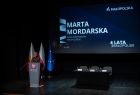 Radna Marta Mordarska w trakcie wystąpienia. Radna stoi przy szarej mównicy. 