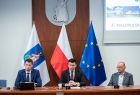 wicemarszałek Łukasz Smółka siedzący na tle flag podczas Forum po obu stronach dwóch innych uczestników obrad